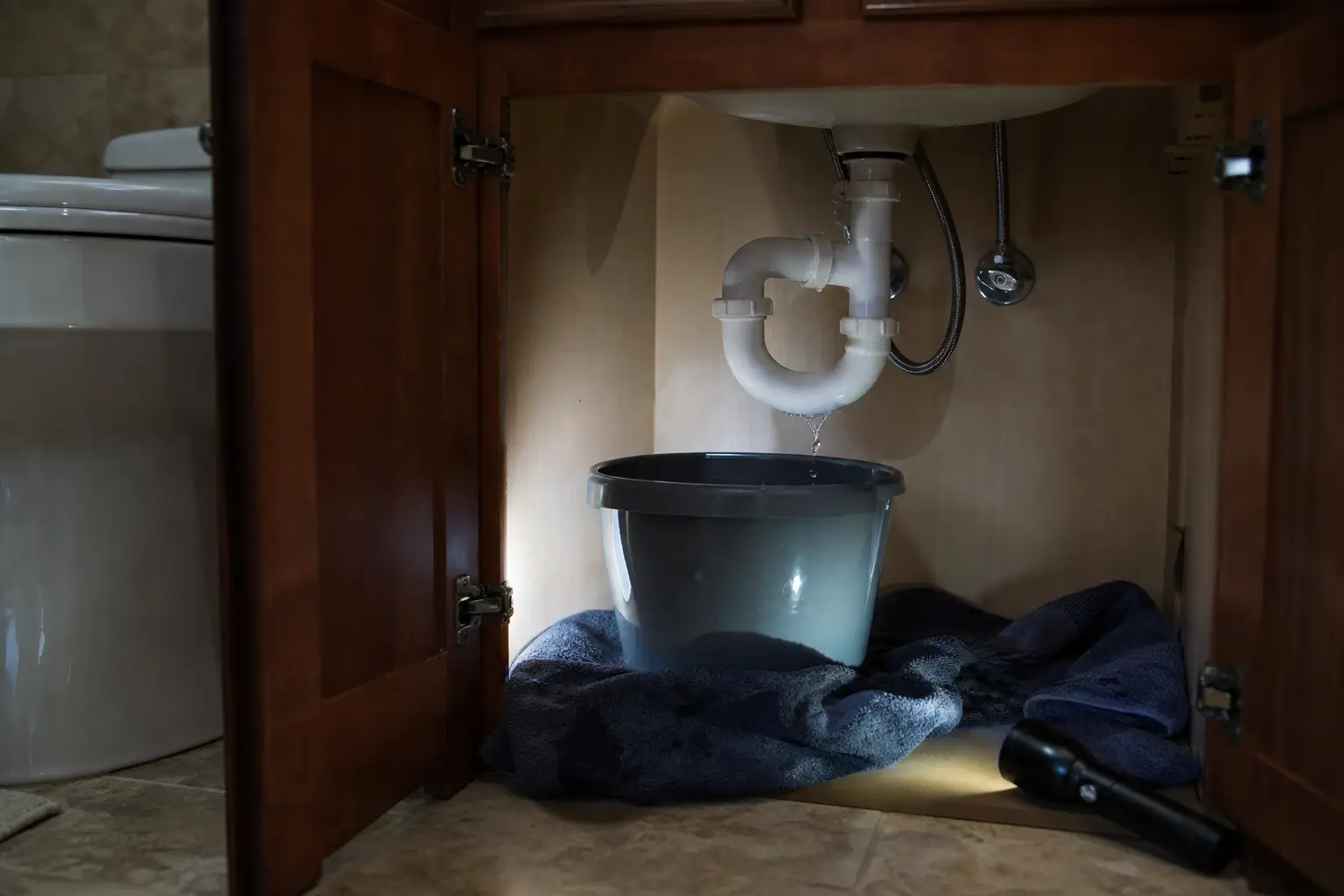 Small persistent leak under a bathroom sink with a towel and bucket catching drips